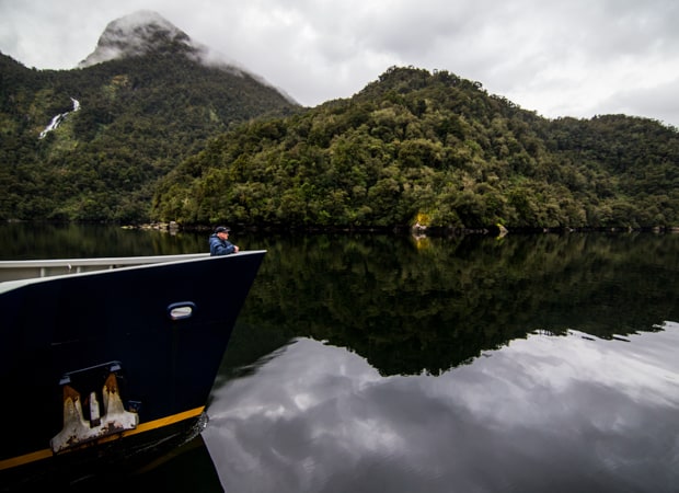 A man in a hat and blue jacket stands at the bow of a catamaran on a New Zealand small ship cruise through Fiordland National Park
