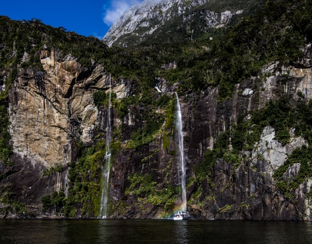 A New Zealand small ship cruise boat is seen under a waterfall and cliff on a sunny day
