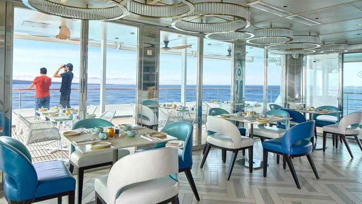 aft indoor/outdoor dining area on Nat Geo Islander II Galapagos ship with airy blue & white tables for 2 & 4 guests.