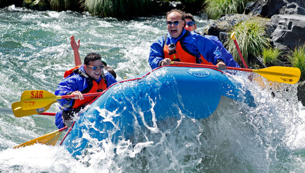 3 men in sunglasses, blue jackets & orange life vests use yellow paddles to move a blue raft through whitewater on a Snake River tour.