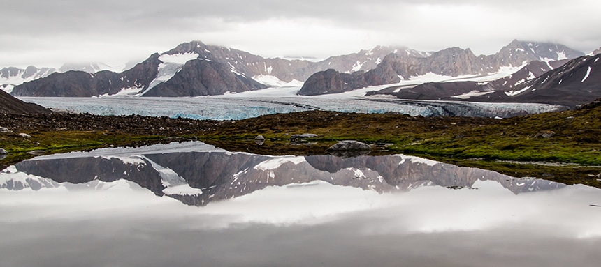 In Arctic Svalbard Norway a massive mile long glacier lays between a mountain range and seen mirrored on a body of water below.