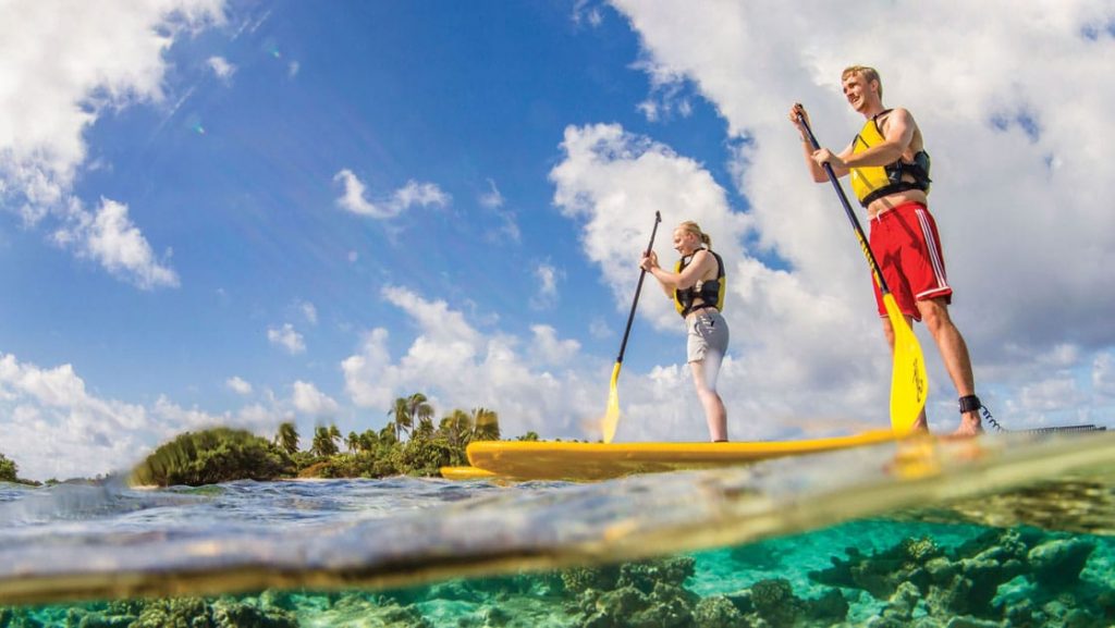 Boy in red swim trunks & girl in tan shorts each paddle a yellow stand-up paddleboard above turquoise water in Fiji.