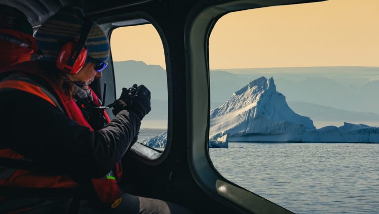 Antarctica traveler photographs a giant iceberg at dusk beyond the window of a helicopter on the Emperor Penguin Quest.