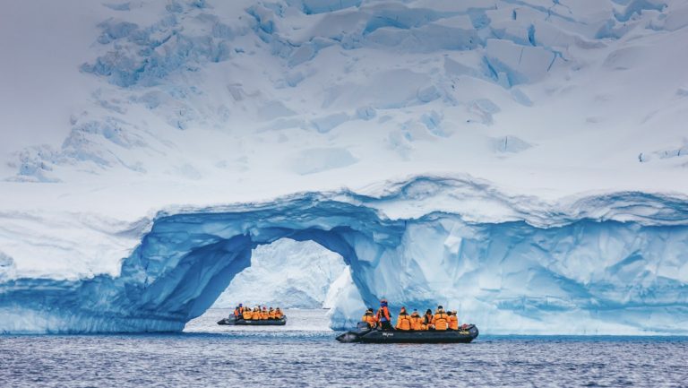 2 zodiacs with Antarctica travelers in bright jackets cruise amongst a snowy arch during an emperor penguin expedition.