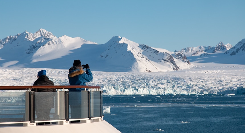 On a bright blue sky day Svalbard cruise guests from the bow of a ship look out over an icy arctic landscape and take photos.