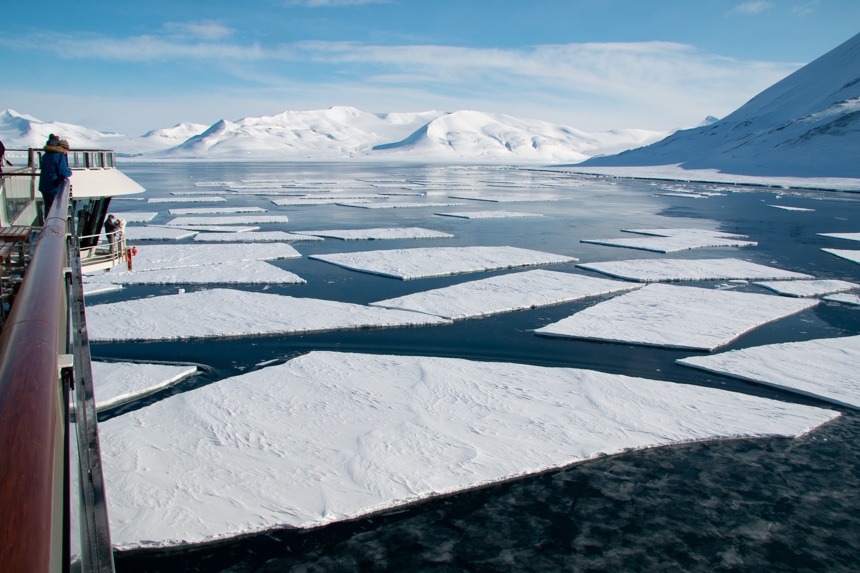 A Svalbard spring landscape of snowy mountain ranges and large white flat ice sheets floating in Arctic waters seen from the ship deck
