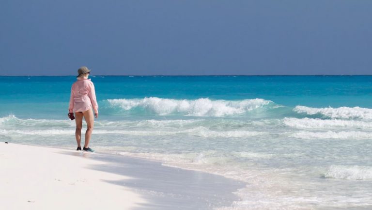 Woman in pink shirt & bathing suit stands on white-sand beach beside turquoise sea during a Bahamas small ship cruise.