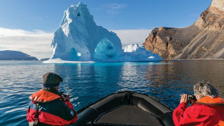 Travelers sit in a Zodiac & photograph a large iceberg beside beige rock on a cruise from Greenland to Canada.