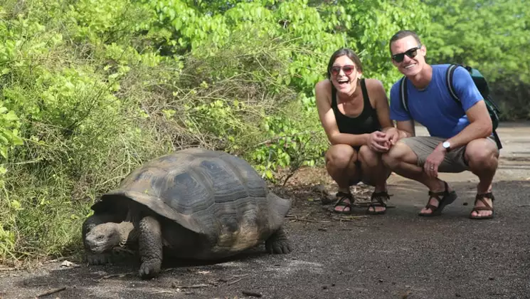Young man & woman kneel down & smile behind a Galapagos tortoise as it walks away during a Grand Majestic cruise.