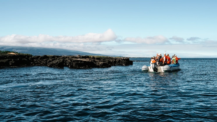 Galapagos travelers ride in a panga boat with an outboard motor, viewing a dark lava rock shore on a Grand Majestic cruise.