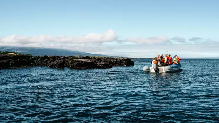 Galapagos travelers ride in a panga boat with an outboard motor, viewing a dark lava rock shore on a Grand Majestic cruise.