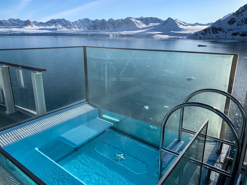 A blue infinity jacuzzi on the Resolution, with glass walls and a scenic arctic landscape view over the stern of the ship.