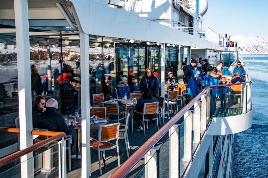 Guests enjoy eating their meal outside at wooden tables on a sunny day in on the top deck of the National Geographic Resolution.