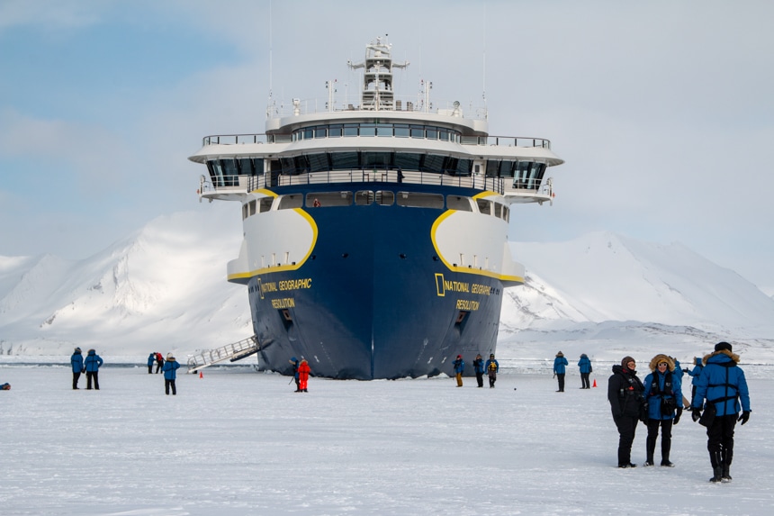 The futuristic blue yellow and white Nat Geo Resolution ship parks in the ice as it's guests walk on the frozen sea around it.