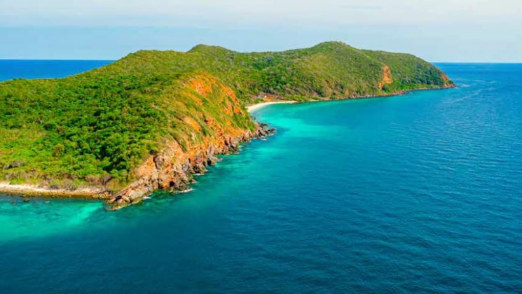 Aerial view of a remote island with lush greenery, red cliffs & turquoise shores seen on the Rites & Relics South Pacific cruise.