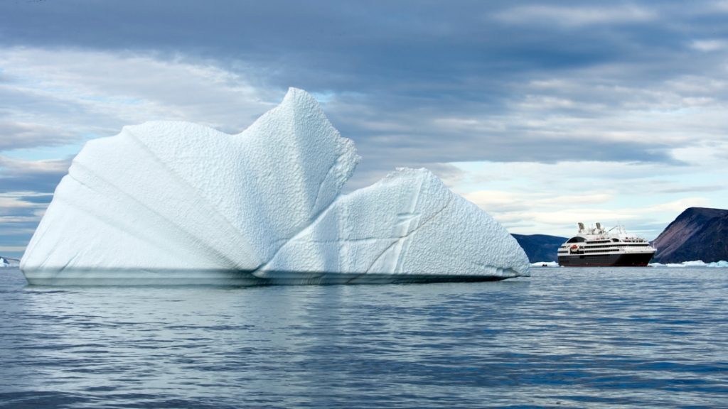 Cruising in Greenland's Baffin Bay is a luxury Ponant ship, dwarfed in size compared to a large iceberg floating next to it.