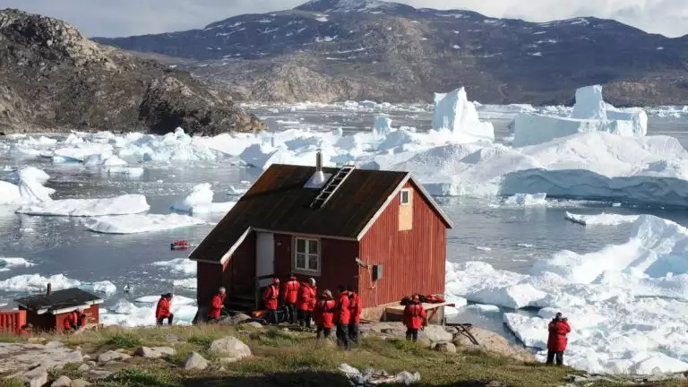 Luxury ponant cruise guests in red parkas explore the shore in Greenland among red wood building and a sea filled with icebergs.