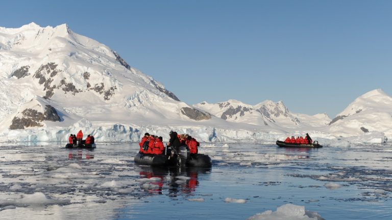 3 black Zodiac boats with polar travelers in red jackets, cruise among ice chunks beside snow-covered hills on a sunny day.