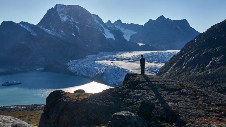 A male guest stands on a rocky hill side overlooking a massive glacier wedged between jagged mountain range that opens to the sea