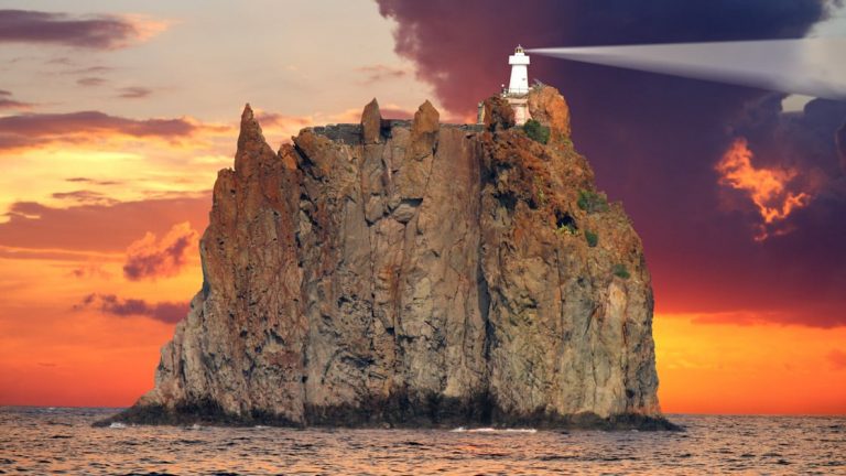 The cliff-top Stromboli lighthouse seen during a bright orange sunset with its lightbeam shining bright off to the right