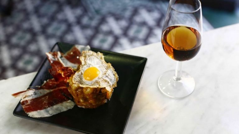 A glass of brown wine seen beside a plate of proscuitto and an egg.