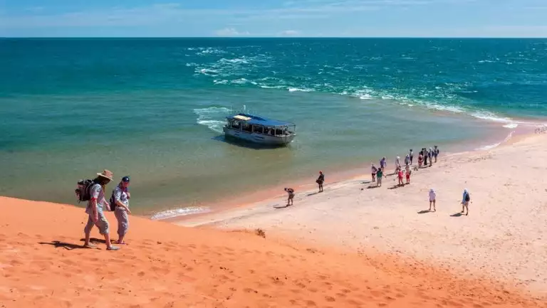 Travelers walk down a red sand hill towards people on the beach as a small aluminum boat pulls into shore in west Australia.