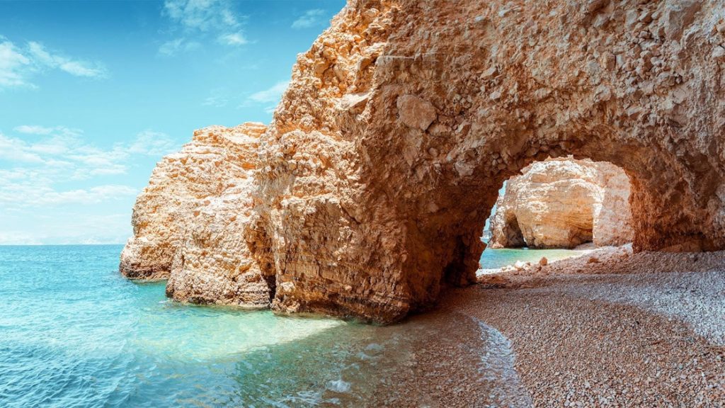 Red seaside cliffs form an arch over the turquoise ocean in Koufonissia, Greece.