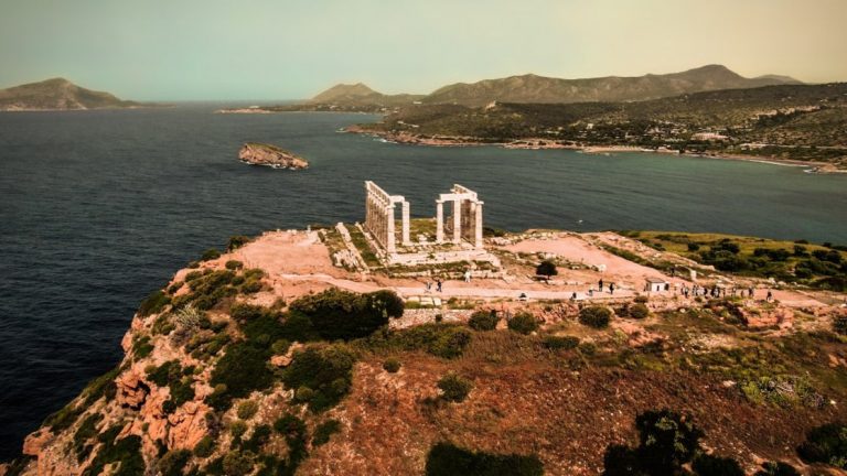 A Greek temple seen from an aerial drone view with ocean and mountains in the background