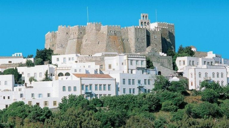 A view of Patmos and its medieval town of Chora behind more modern white buildings and green trees.
