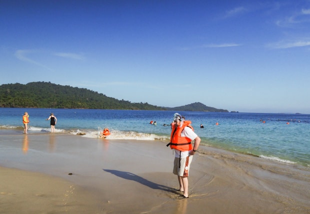 Traveler in a orange life vest on the beach after a snorkel with other travelers frolicking on the beach and water.