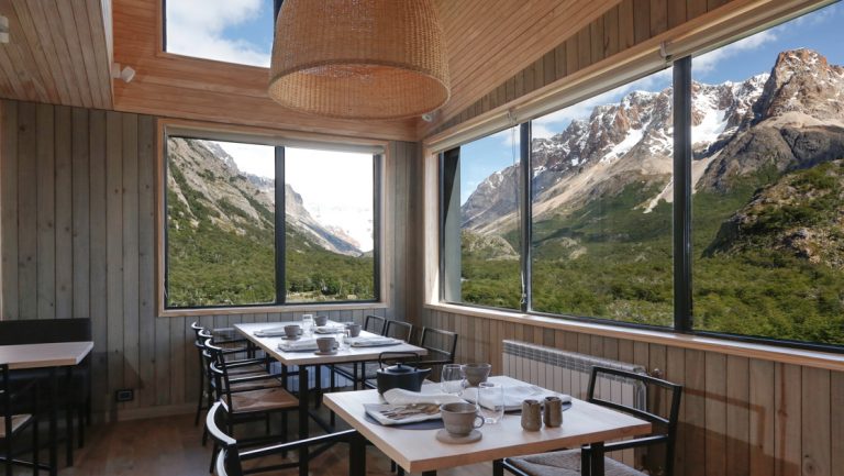 Dining room at Explora Hotel El Chalten with wood 4-top tables, metal chairs, wall-to-wall windows & skylights onto mountains.