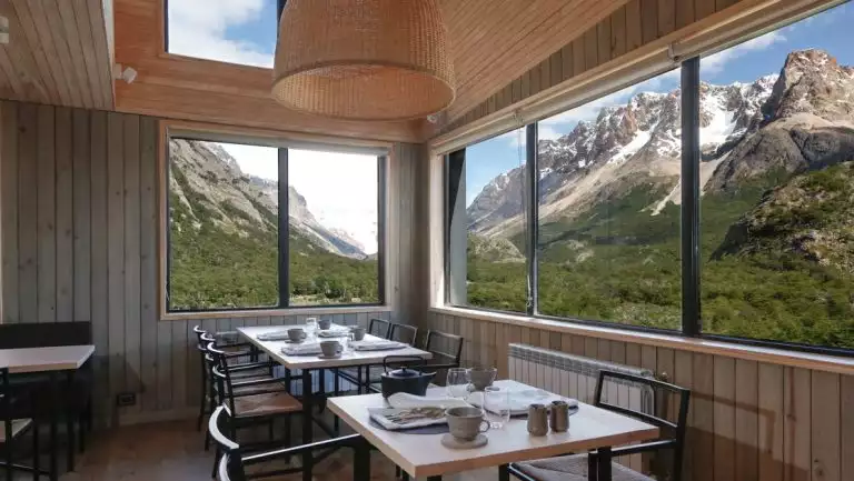 Dining room at Explora Hotel El Chalten with wood 4-top tables, metal chairs, wall-to-wall windows & skylights onto mountains.