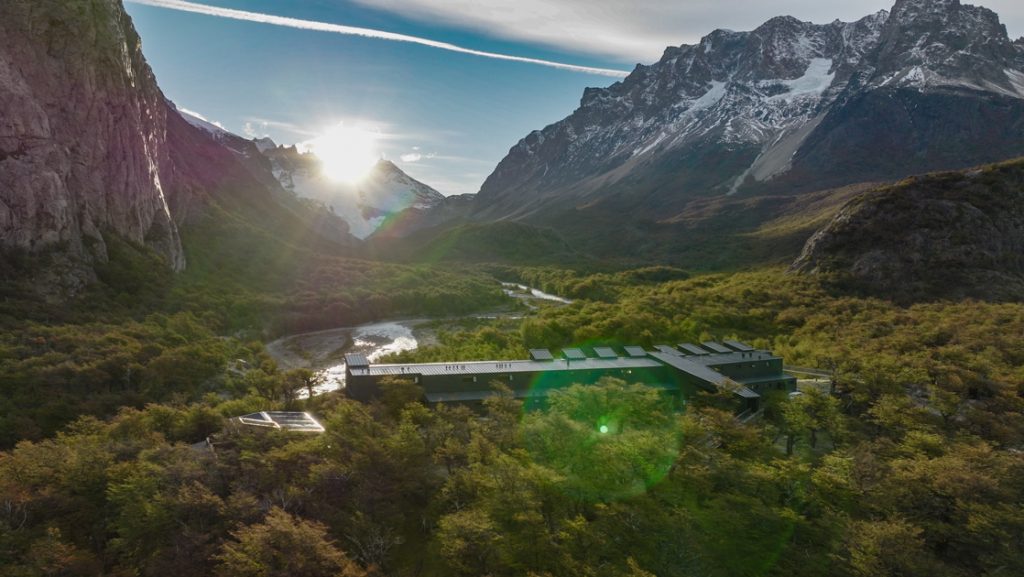 Aerial view of a dark metal, rectangular hotel sitting beside a river in a valley of trees by tall, snow-capped peaks.