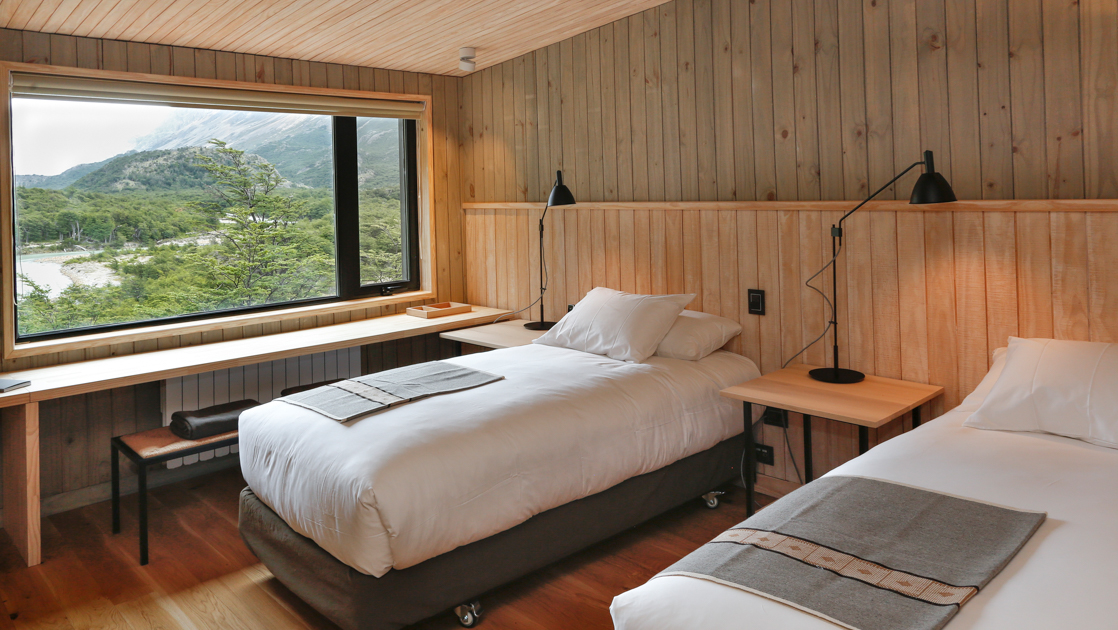2 twin beds against wood wall beside view windows & desk in earth tones decor at Explora Lodge El Chalten.