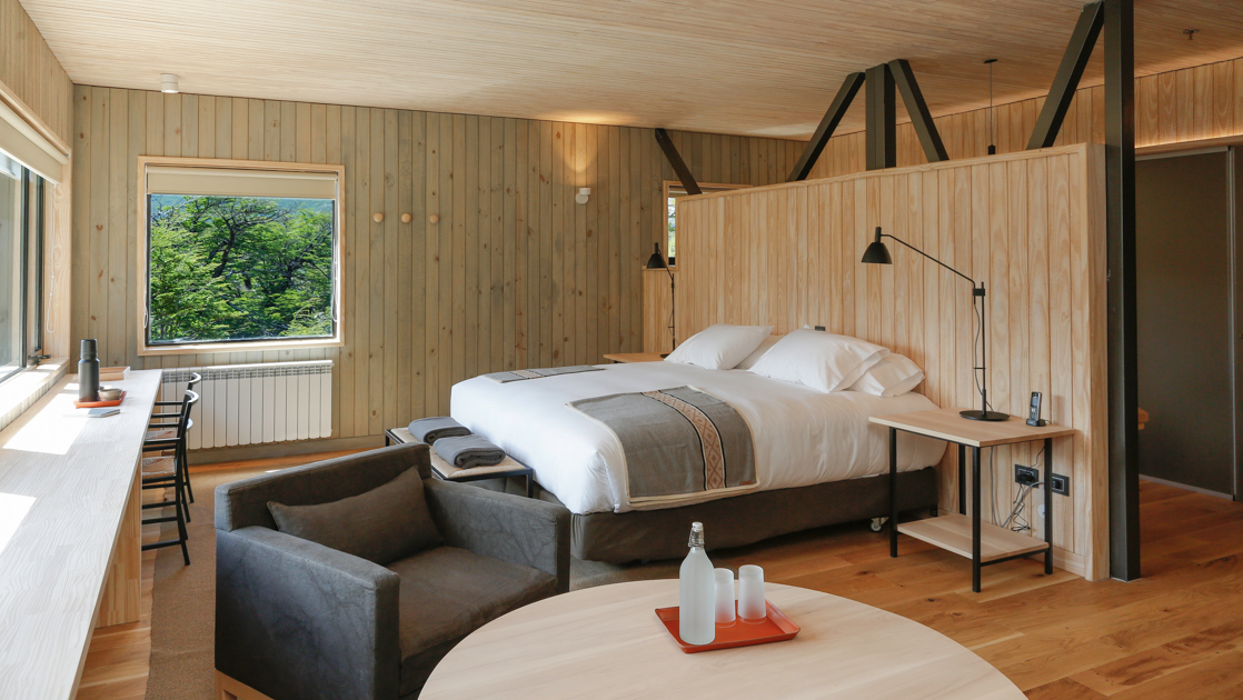 King bed with wood half-wall behind & view windows & seating area in front in earthtones decor at Explora Lodge El Chalten.