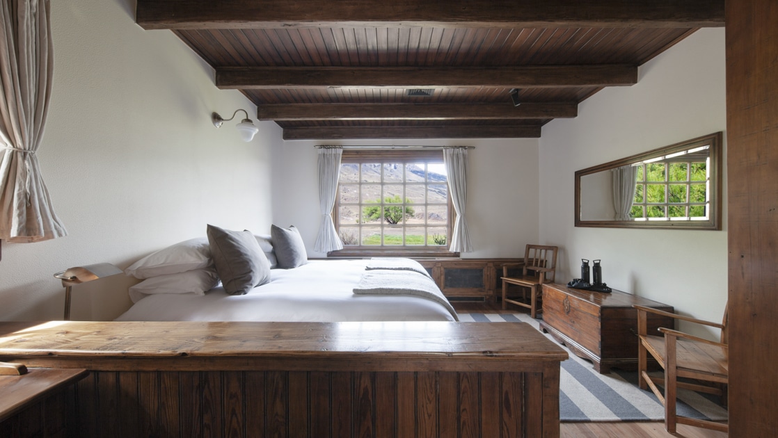 Room with large bed in white linens by wood furniture & ceiling & large latticed window looking out to rugged mountains.