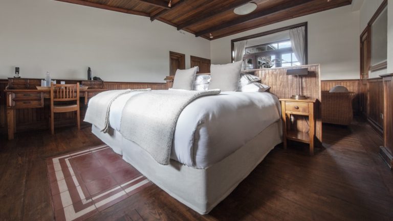 Double bed in white linens sits in glow from the window with wood furniture, floor & ceiling beams at Explora Patagonia NP Lodge.