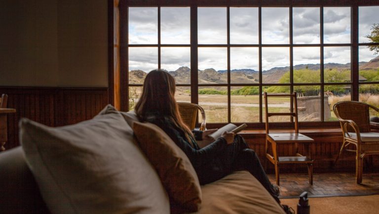 Woman sits & reads facing large window looking out onto Patagonia National Park's grasslands with mountains in the distance.