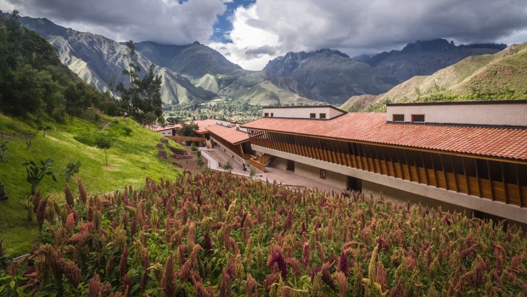 Looking down on the long, stone & wood Explora Sacred Valley lodge in a bright green Andean valley with purple flowers.