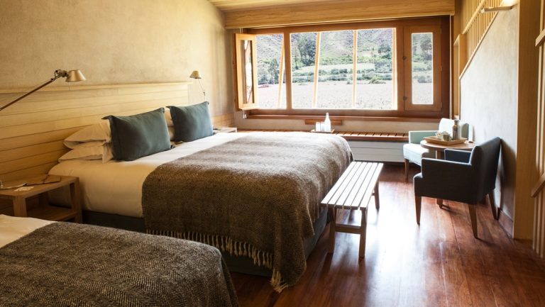 Room at Explora Sacred Valley lodge with 2 beds, large window, table & 2 chairs in a minimalist decor bathed in natural light.