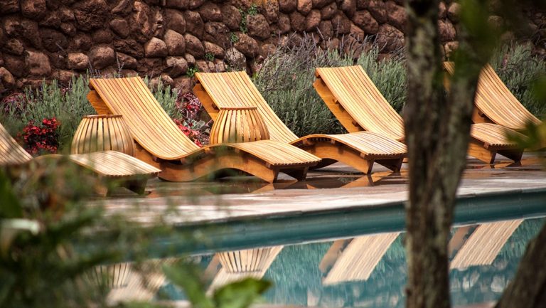 Empty wood chaise lounge chairs with basket-like side tables sit against a stone wall by a pool at Explora Sacred Valley Lodge.