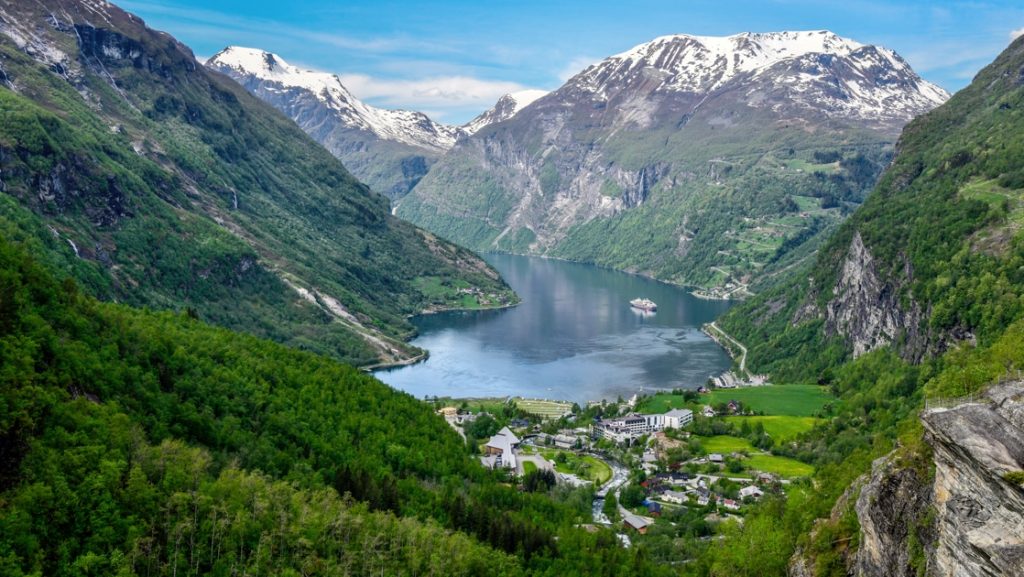 Aerial view of lush green mountains with rocky, snowy tops leading down to a small town beside a calm body of water with a ship.