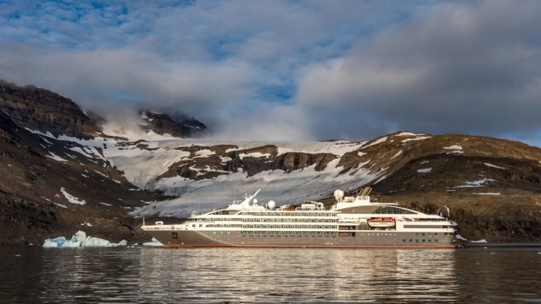 The luxury Ponant ship, painted grey and white with a red stripe float along a partly snowy mountainous coastline of Greenland.
