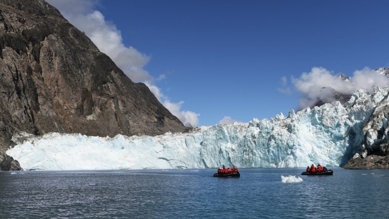At the end of a fjord in Greenland two groups take a skiff riding activity and cruise in front of a icy teal glacier.