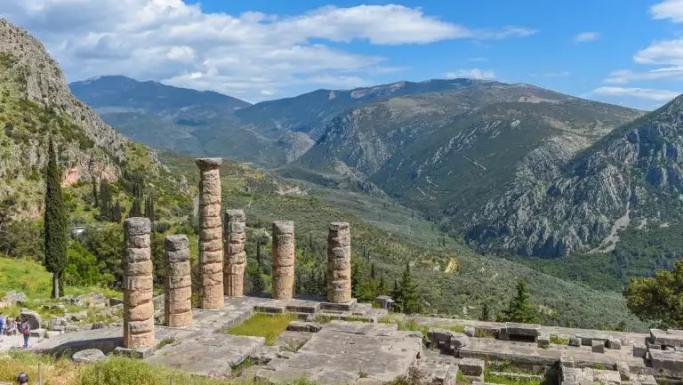 Looking down on a row of ancient stone pillars sitting among ruins atop a hillside on a sunny day in Greece.