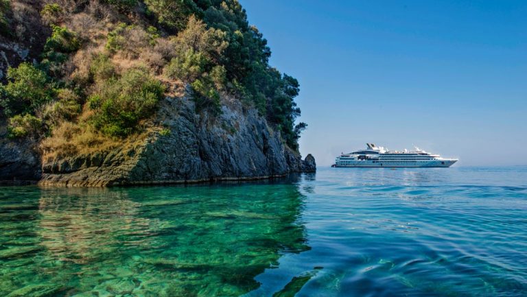 Water-level view of a small island beside clear green & blue sea with a small expedition ship in the background.