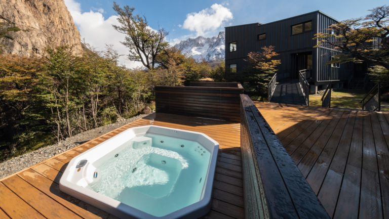 Empty Jacuzzi ringed by low wood walls sits beside a dark hotel building, forest & tall mountains at Explora Lodge El Chalten.