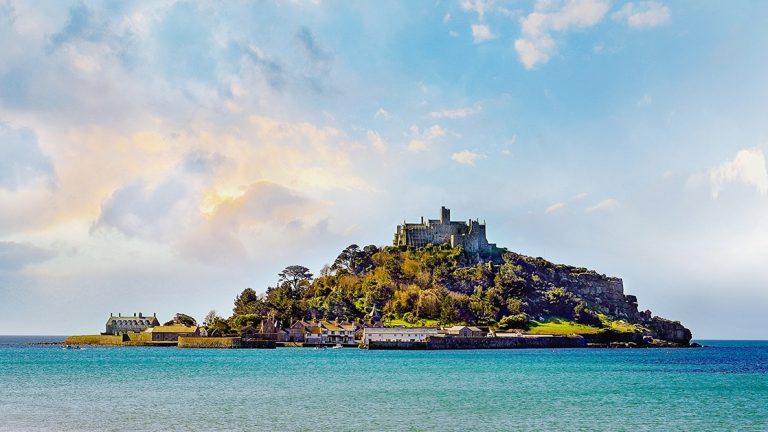 A from-the-water view of St. Michaels Mount in Penzance, Cornwall, seen with bright blue water and a sunny sky with wispy clouds lit by the sun.