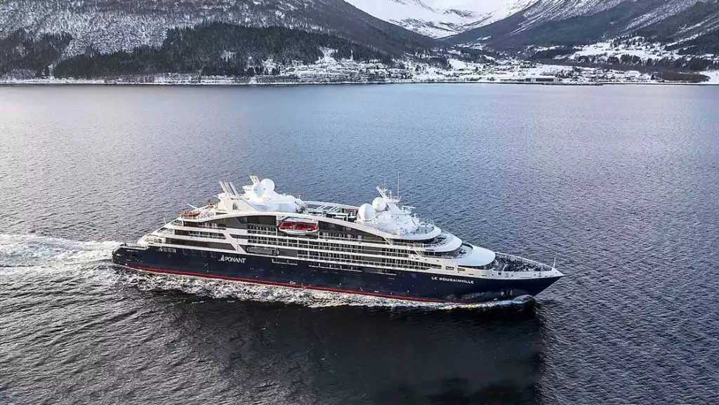 Le Bougainville cruise ship with blue hull, curved white upper decks & red accents sails past snowy mountains.