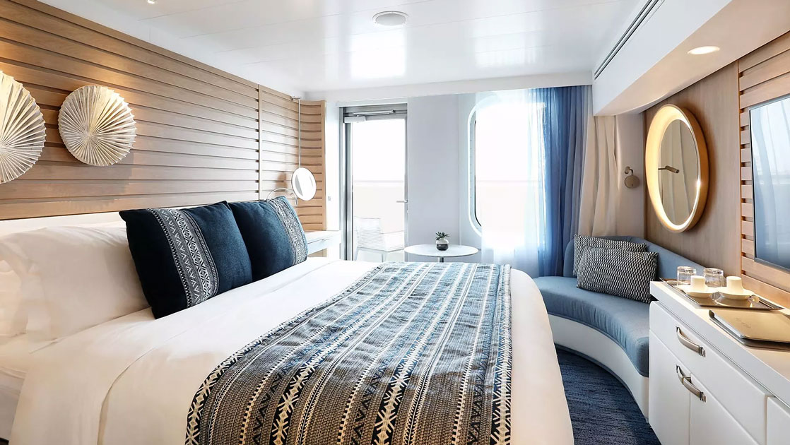 View from bed to dresser & couch beside window & door to balcony in the blue-accented Deluxe Stateroom on Le Bougainville ship.